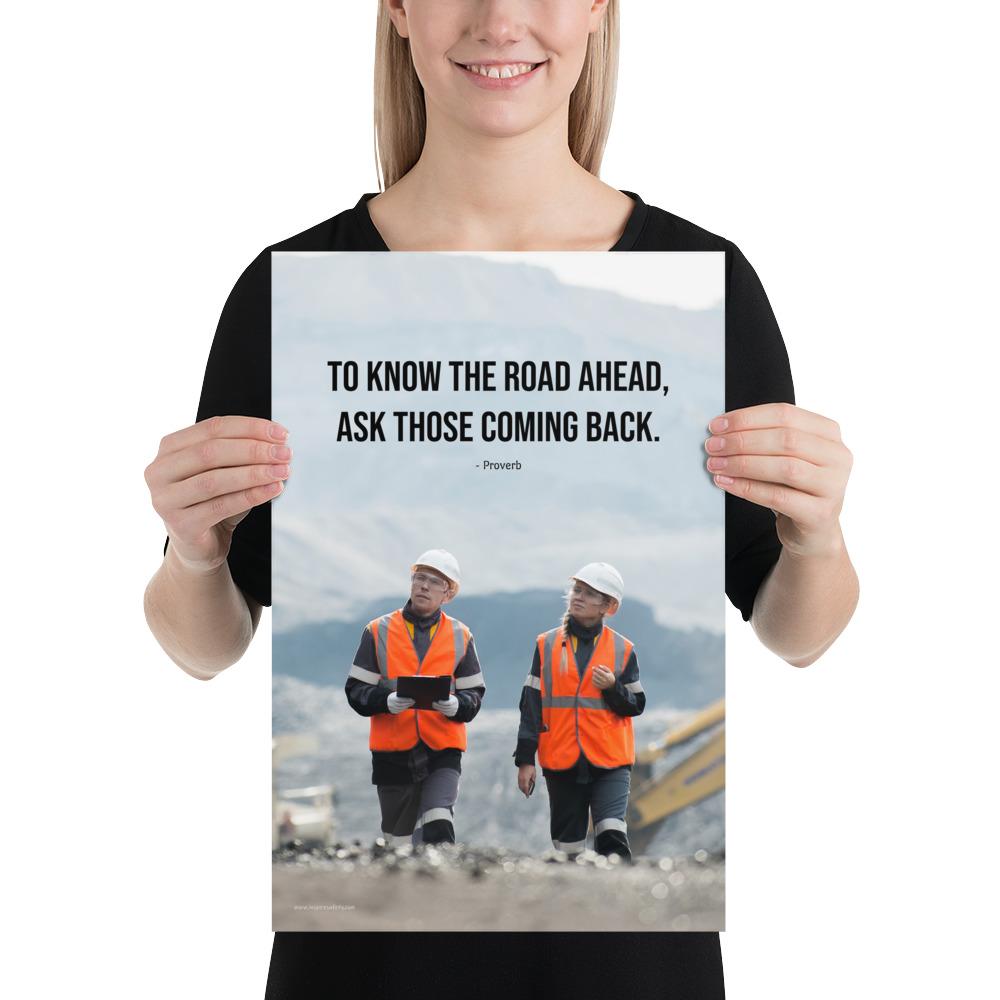 A safety poster showing two workers in reflective vests collaborating on a construction site with the proverb to know the road ahead ask those coming back.