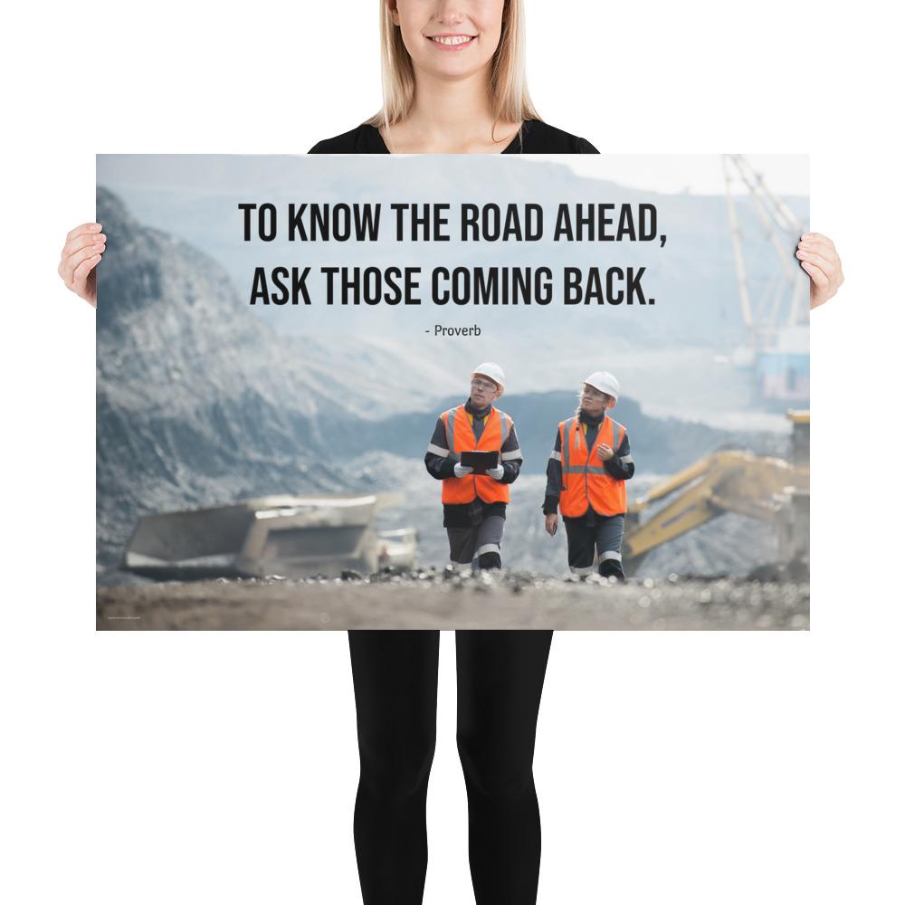 A safety poster showing two workers in reflective vests collaborating on a construction site with the proverb to know the road ahead ask those coming back.