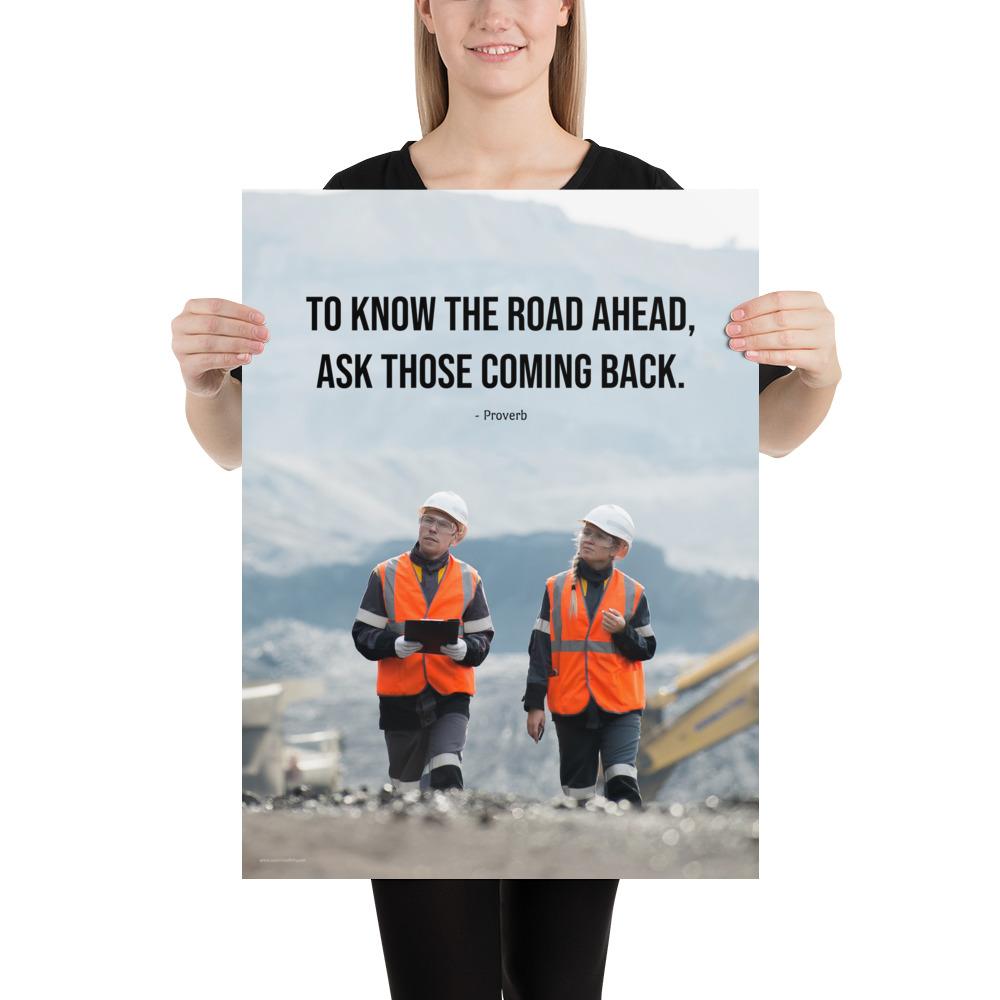 A safety poster showing two workers in reflective vests collaborating on a construction site with the proverb to know the road ahead ask those coming back.