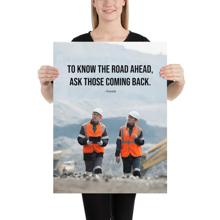 A safety poster showing two workers in reflective vests collaborating on a construction site with the proverb to know the road ahead ask those coming back.