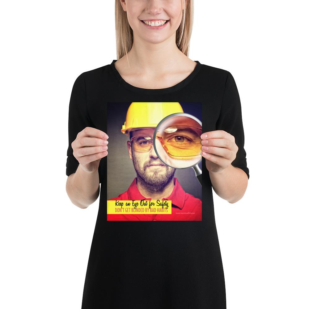 A workplace safety poster showing a portrait of a man wearing a hardhat and safety glasses with a magnifying glass magnifying one eye with the slogan, keep an eye out for safety, don't get blinded by bad habits.