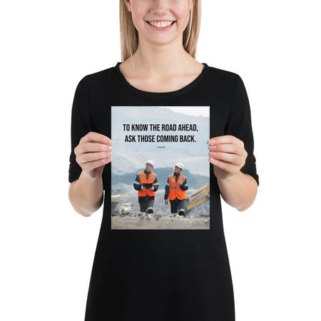 A safety poster showing two workers in reflective vests collaborating on a construction site with the proverb to know the road ahead ask those coming back.