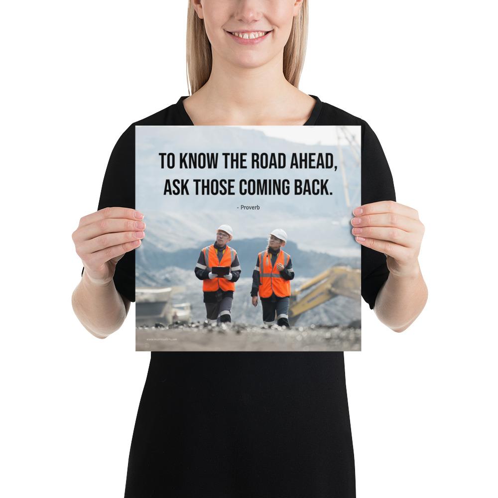A safety poster showing two workers in reflective vests collaborating on a construction site with the proverb to know the road ahead ask those coming back.