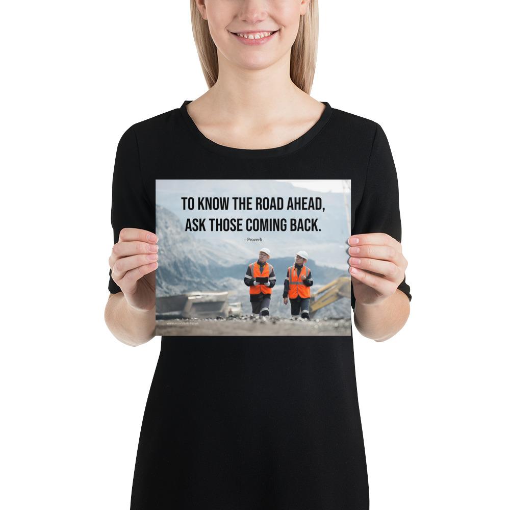 A safety poster showing two workers in reflective vests collaborating on a construction site with the proverb to know the road ahead ask those coming back.