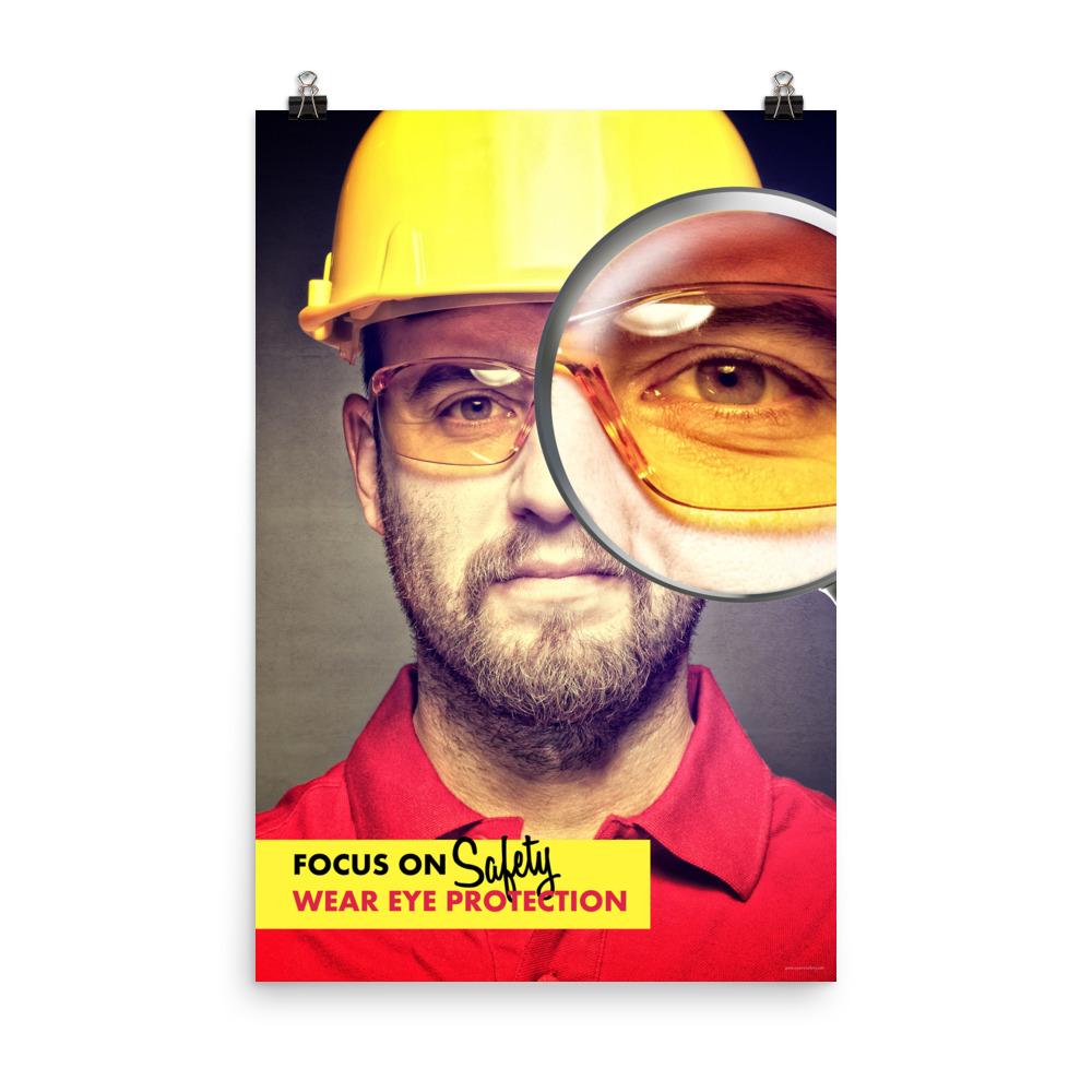 An eye safety poster showing a close up of a man's face wearing safety glasses and a yellow hard hat with a magnifying glass focusing on the right eye with an eye safety slogan underneath him.