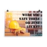 A workplace safety poster showing a yellow hard hat and some gloves sitting on a brick wall on a construction site with the slogan were you safe today, or just lucky written in black stencil font.