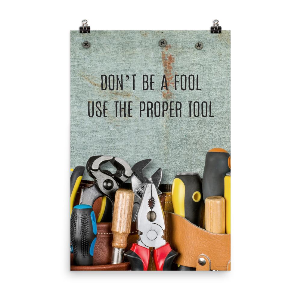 A workplace safety poster showing various tools such as a screwdriver, wrench, and pliers on a wooden table with the slogan don't be a fool use the proper tool.