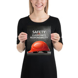 A workplace safety poster showing a red hard hat in front of a grey, industrial-looking background with the slogan safety is everyone's responsibility.