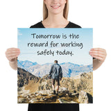 A workplace safety poster showing a man standing on a peak of a mountain with a bright blue sky and mountains behind him with the slogan tomorrow is the reward for working safely today.