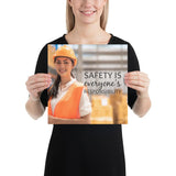 A workplace safety poster showing a young warehouse worker in a yellow hardhat and orange reflective vest holding a clipboard and smiling with the slogan safety is everyone's responsibility.