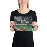 A workplace safety poster depicting a beautiful landscape of mountains in the background of a lake with a little wooden boat in the lake with text saying live safely work safely.