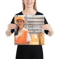 A workplace safety poster showing a young warehouse worker in a yellow hardhat and orange reflective vest holding a clipboard and smiling with the slogan safety is everyone's responsibility.