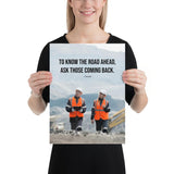 A safety poster showing two workers in reflective vests collaborating on a construction site with the proverb to know the road ahead ask those coming back.
