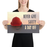 A workplace safety poster depicting a red hard hat sitting on a concrete wall with a dreamy pink sunset in the background with the text never give safety a day off to the right.