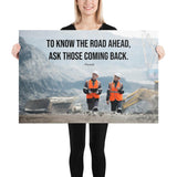 A safety poster showing two workers in reflective vests collaborating on a construction site with the proverb to know the road ahead ask those coming back.