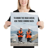 A safety poster showing two workers in reflective vests collaborating on a construction site with the proverb to know the road ahead ask those coming back.