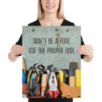 A workplace safety poster showing various tools such as a screwdriver, wrench, and pliers on a wooden table with the slogan don't be a fool use the proper tool.