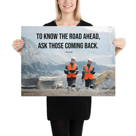 A safety poster showing two workers in reflective vests collaborating on a construction site with the proverb to know the road ahead ask those coming back.
