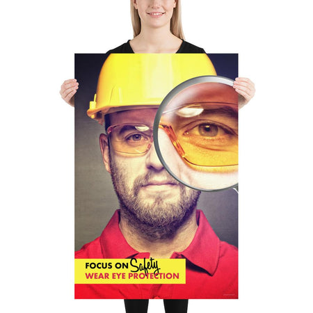 An eye safety poster showing a close up of a man's face wearing safety glasses and a yellow hard hat with a magnifying glass focusing on the right eye with an eye safety slogan underneath him.