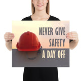 A workplace safety poster depicting a red hard hat sitting on a concrete wall with a dreamy pink sunset in the background with the text never give safety a day off to the right.