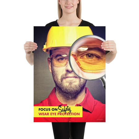 An eye safety poster showing a close up of a man's face wearing safety glasses and a yellow hard hat with a magnifying glass focusing on the right eye with an eye safety slogan underneath him.