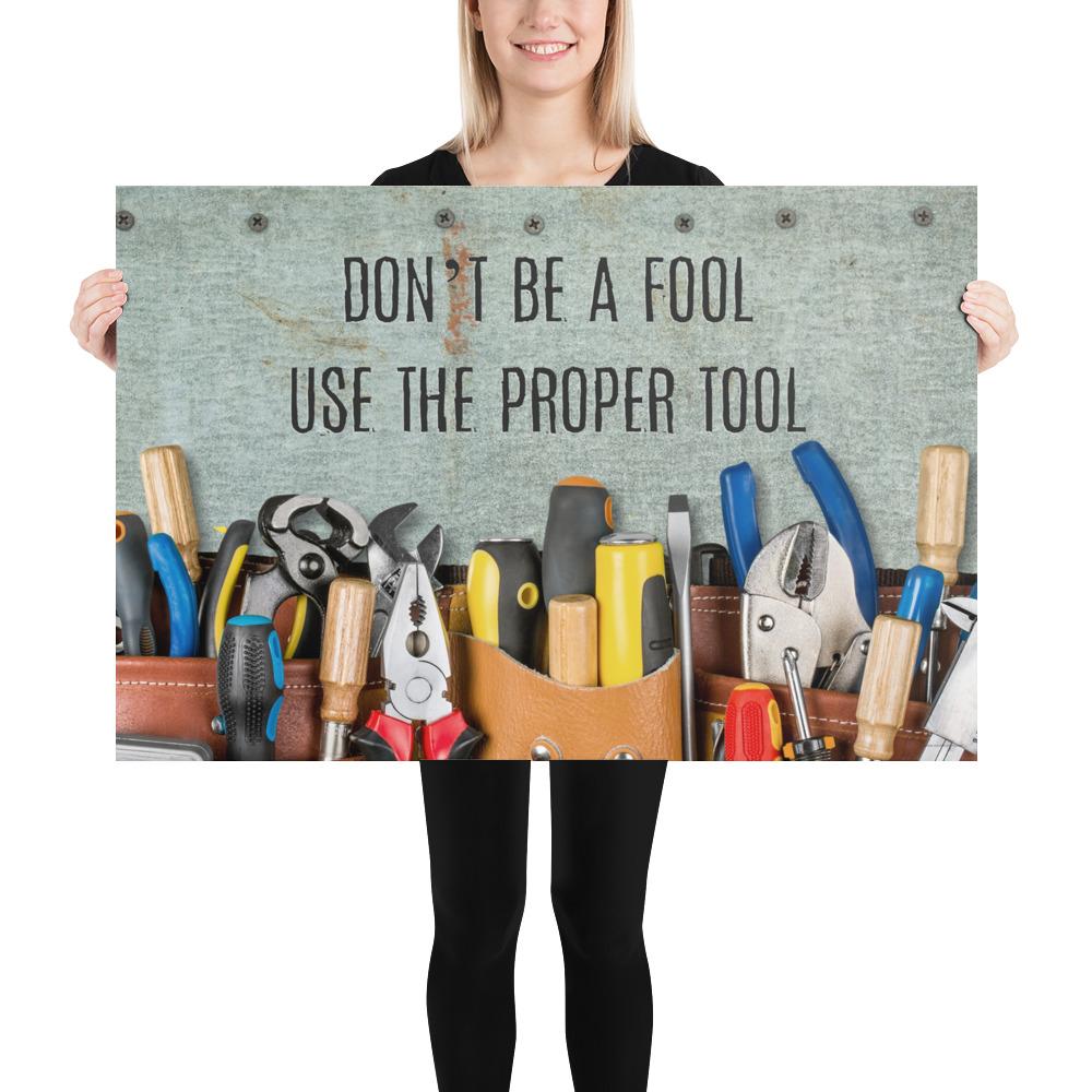 A workplace safety poster showing various tools such as a screwdriver, wrench, and pliers on a wooden table with the slogan don't be a fool use the proper tool.