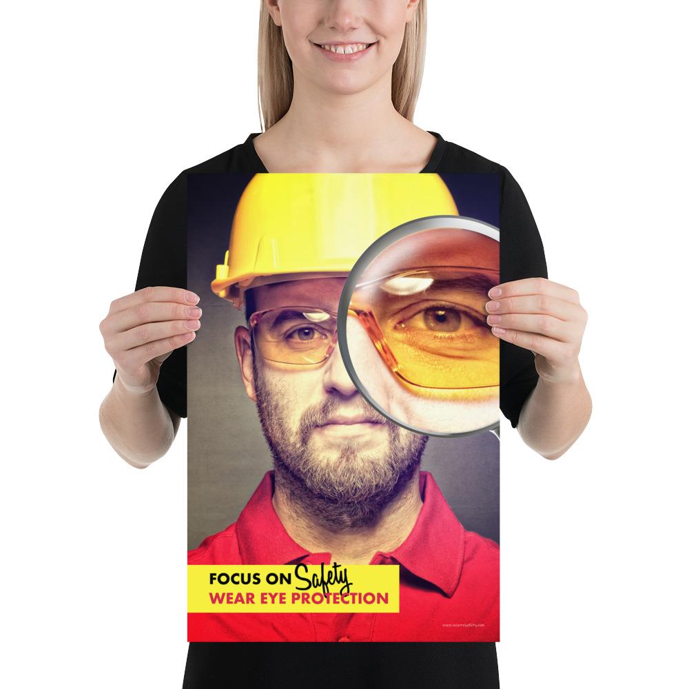 An eye safety poster showing a close up of a man's face wearing safety glasses and a yellow hard hat with a magnifying glass focusing on the right eye with an eye safety slogan underneath him.