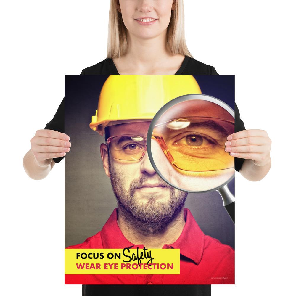 An eye safety poster showing a close up of a man's face wearing safety glasses and a yellow hard hat with a magnifying glass focusing on the right eye with an eye safety slogan underneath him.