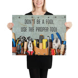 A workplace safety poster showing various tools such as a screwdriver, wrench, and pliers on a wooden table with the slogan don't be a fool use the proper tool.