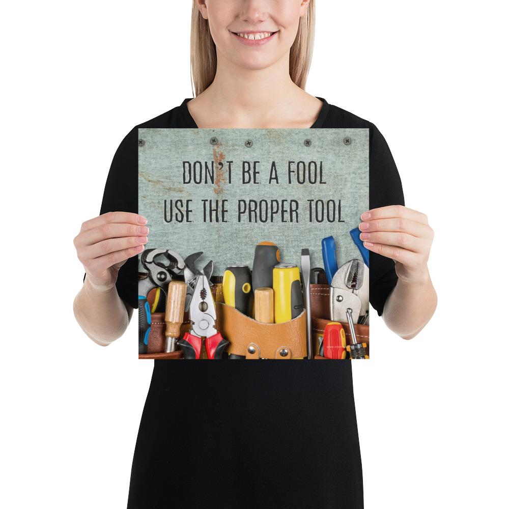 A workplace safety poster showing various tools such as a screwdriver, wrench, and pliers on a wooden table with the slogan don't be a fool use the proper tool.