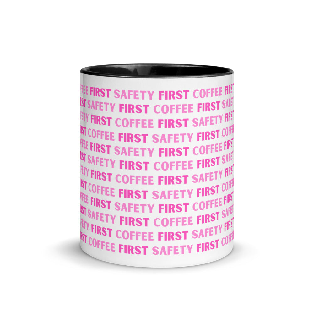 White ceramic mug with pink repeating text that says "Safety First, Coffee First" with a black rim, inside, and handle.