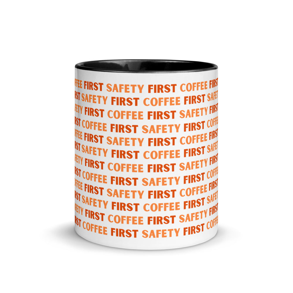 White ceramic mug with orange repeating text that says "Safety First, Coffee First" with a black rim, inside, and handle.