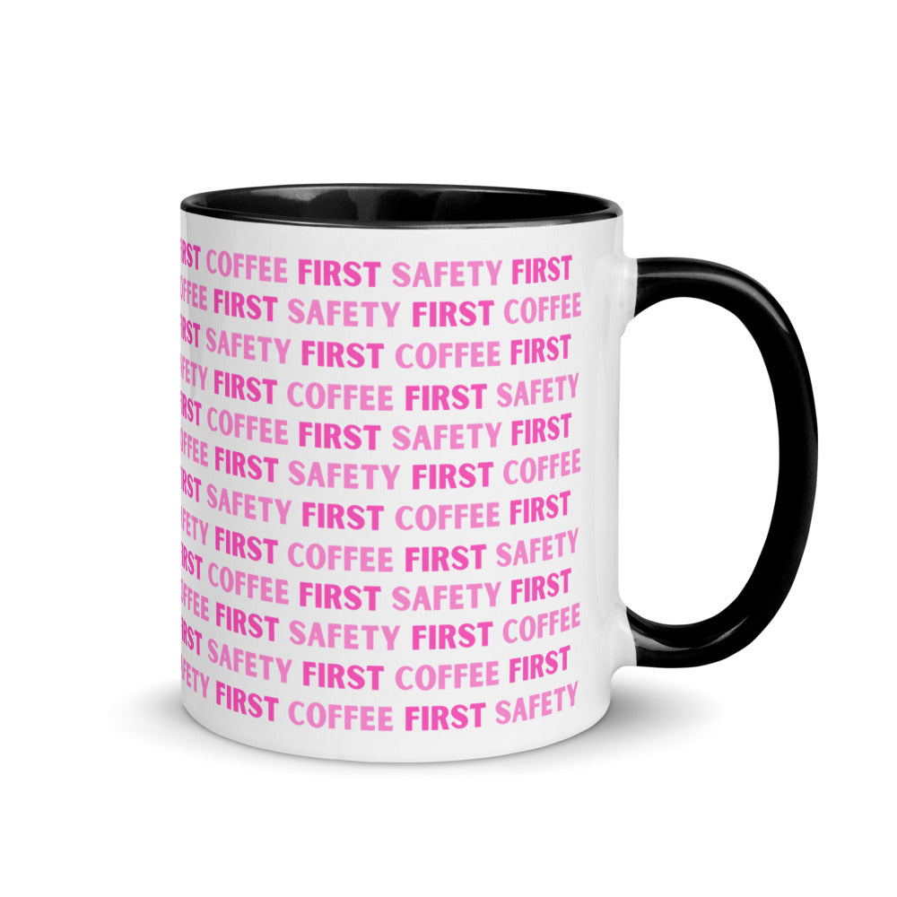 White ceramic mug with pink repeating text that says "Safety First, Coffee First" with a black rim, inside, and handle.